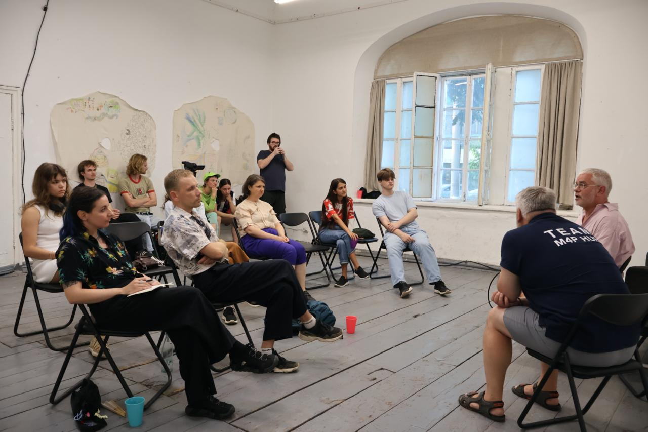 an audience of about a dozen people is facing two presenters in an art space